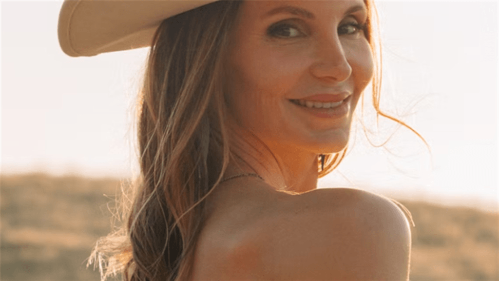A woman with long hair smiles warmly while wearing a cowboy hat in a golden field