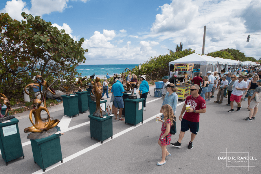 A lively outdoor art festival with bronze sculptures on display along a coastal road, where visitors stroll and enjoy the ocean view