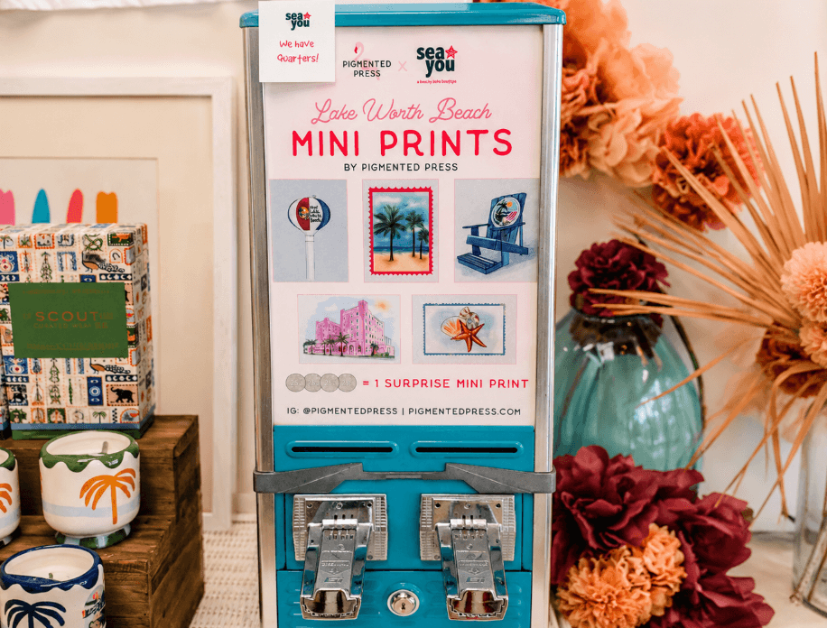 A vibrant display featuring a mini print vending machine with colorful beach-themed artwork, surrounded by decorative flowers and tropical-themed items