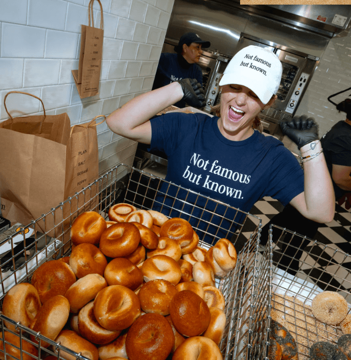 A worker in a bakery proudly displays a large basket of freshly baked bagels, wearing a shirt that reads 'Not famous but known'