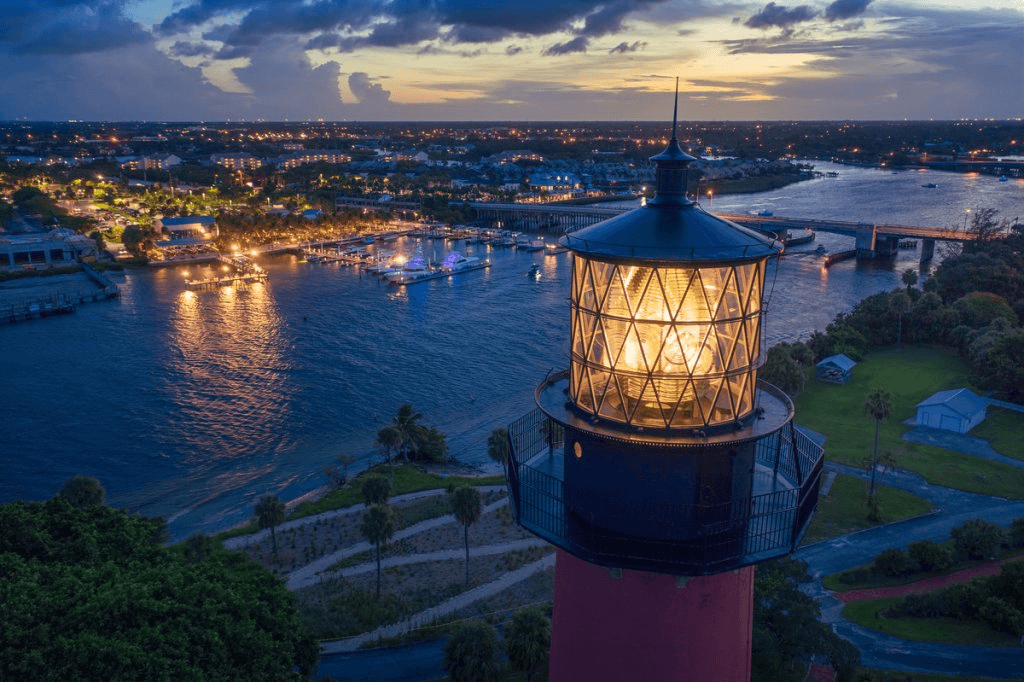 A picturesque coastal scene at dusk, featuring a towering lighthouse with a glowing lantern room, overlooking a serene harbor filled with boats and a vibrant cityscape illuminated by countless lights