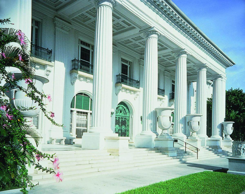 A grand white building with tall columns and a green door, surrounded by lush greenery and pink flowers