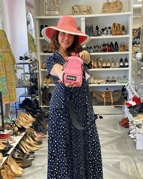 Uma mulher com um vestido de bolinhas e chapéu de abas largas segura uma mochila rosa em miniatura em uma loja de calçados