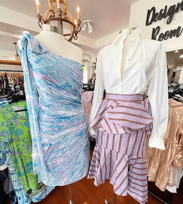 Duas manequins exibem vestidos elegantes: uma com um vestido de um ombro só em tom pastel e a outra com uma blusa branca com uma saia listrada com babados