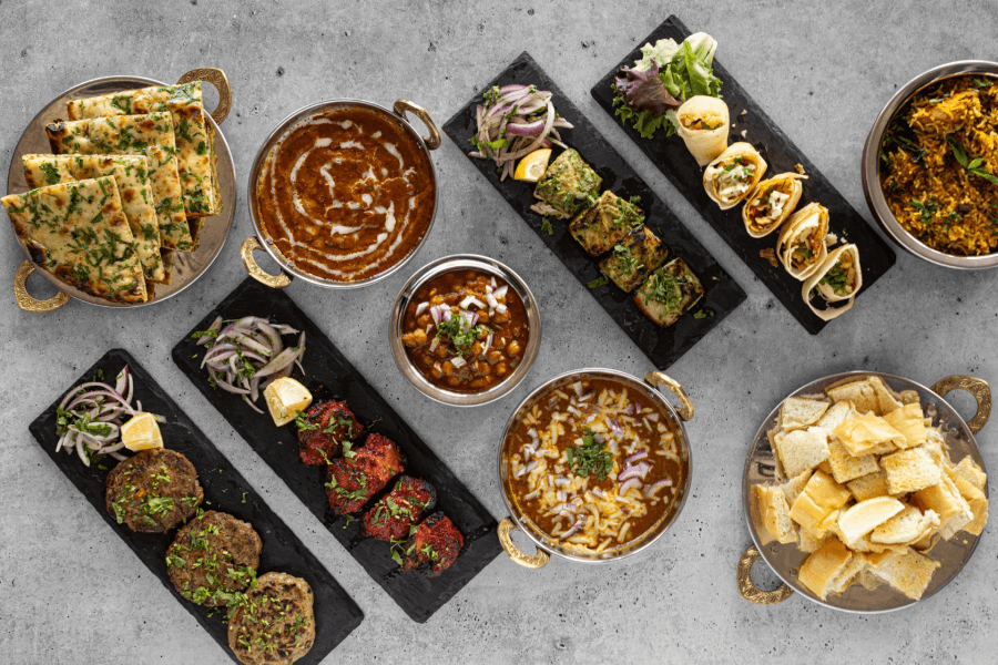 A vibrant spread of Indian dishes including naan bread, curries, kebabs, biryani, and samosas, all artfully arranged on a concrete table
