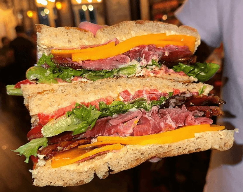 A hand holding a large, layered sandwich with meat, cheese, and lettuce on bread