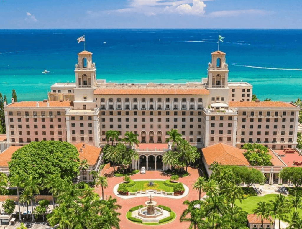 Un majestuoso hotel histórico con dos torres y un tejado de tejas rojas se alza en medio de exuberantes jardines tropicales, con vistas a un vibrante océano de color turquesa