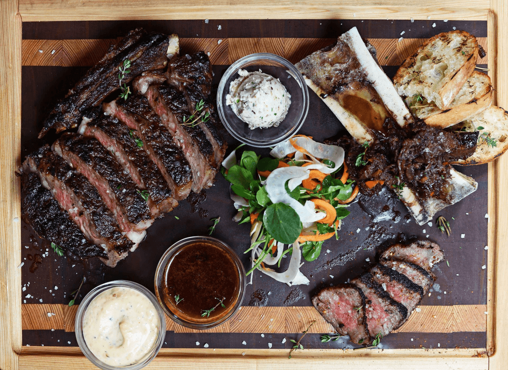 A mouthwatering charcuterie board featuring a variety of meats, including a perfectly cooked ribeye steak, beef short ribs, and slices of prosciutto, accompanied by an assortment of sauces, fresh vegetables, and crusty bread