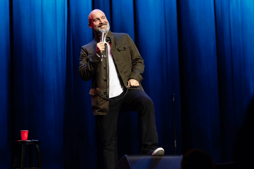Um homem careca com barba, vestindo uma jaqueta verde sobre uma camisa branca, está confiante no palco segurando um microfone, pronto para fazer stand-up comedy