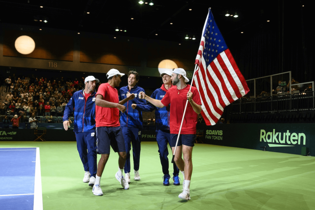 Cinco atletas comemoram sua vitória em uma quadra de tênis, segurando uma bandeira americana no alto