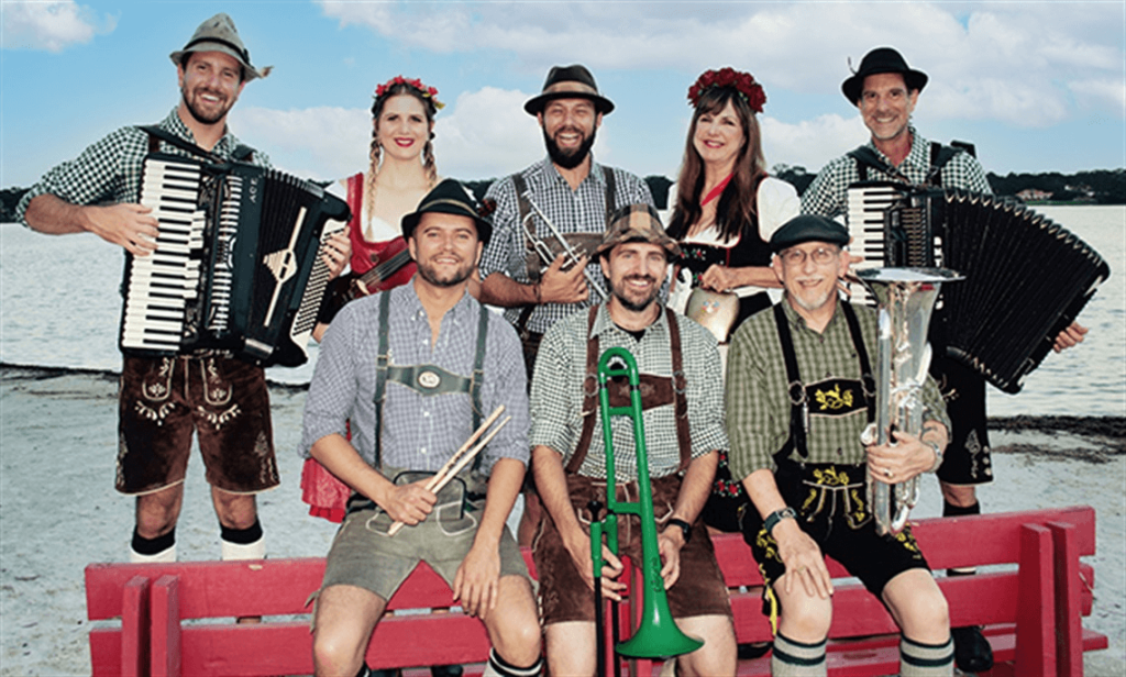 Um animado grupo de músicos vestidos com trajes tradicionais da Baviera, incluindo lederhosen e dirndls, posam juntos em um banco vermelho à beira de um lago, tocando instrumentos como acordeões, tuba e trompete