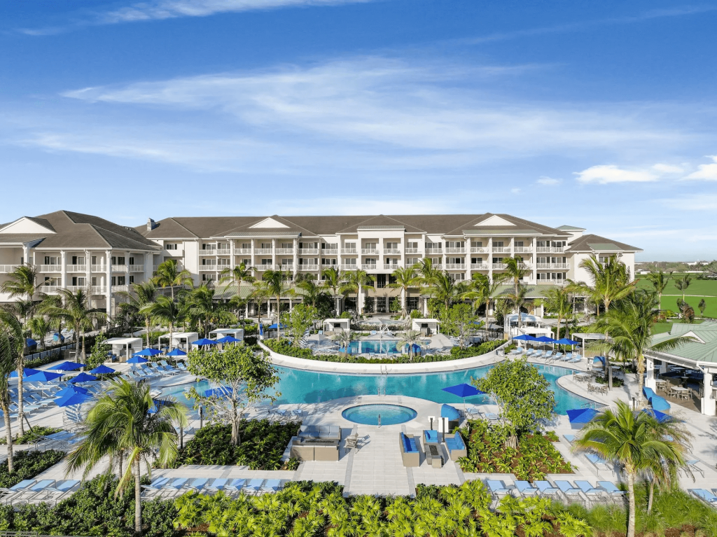 Um luxuoso resort tropical com uma grande piscina sinuosa cercada por palmeiras, espreguiçadeiras e guarda-sóis azuis, tendo como pano de fundo um edifício branco de vários andares com varandas e um céu azul claro