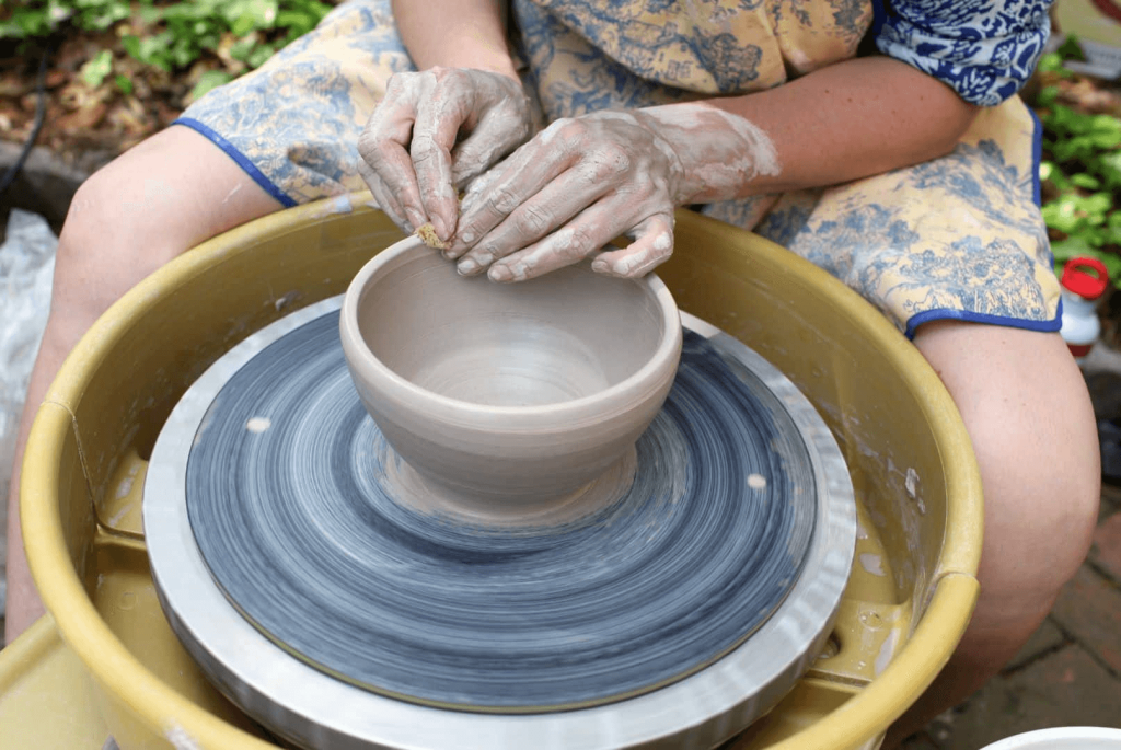 Una persona que lleva un delantal amarillo con motivos florales azules está dando forma con destreza a una vasija de arcilla en un torno de alfarero, con las manos cubiertas de arcilla húmeda