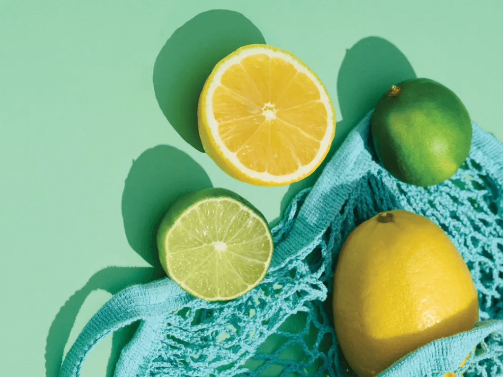 Uma exibição vibrante de frutas cítricas, incluindo um limão amarelo cortado ao meio, um limão inteiro e um limão fatiado, todos descansando em uma bolsa de malha azul brilhante contra um fundo verde refrescante