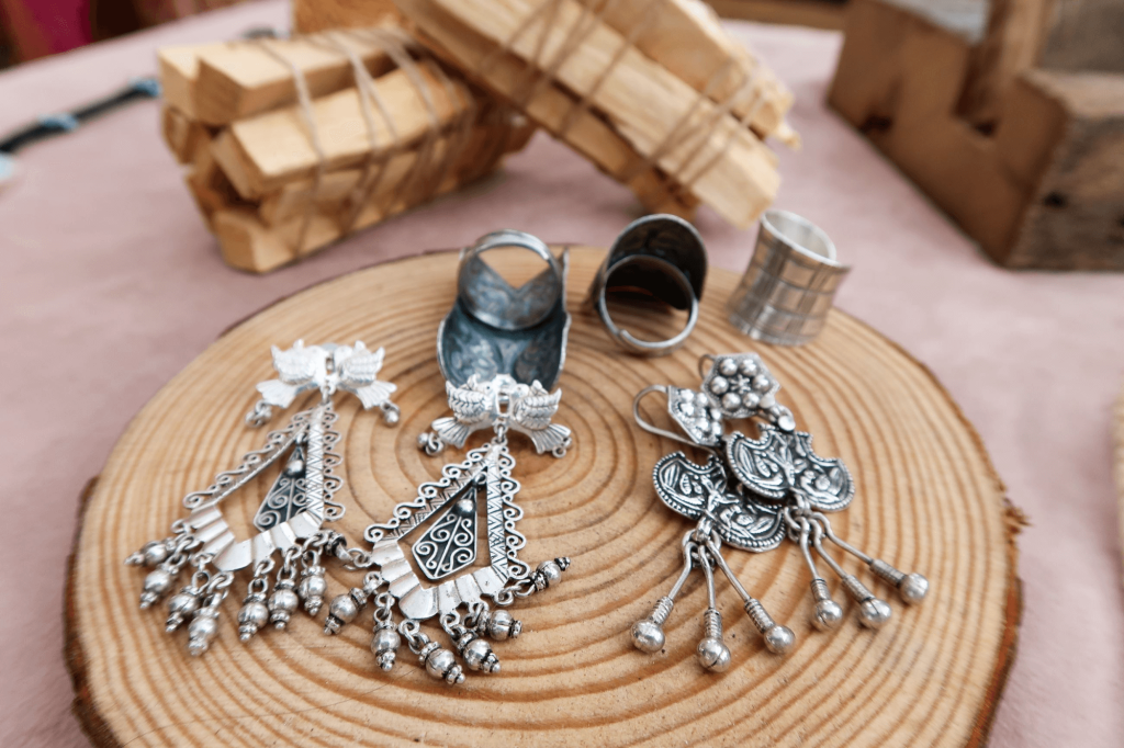 Joias de prata complexas exibidas em uma fatia de madeira, incluindo brincos, anéis e pulseiras com filigranas e miçangas detalhadas