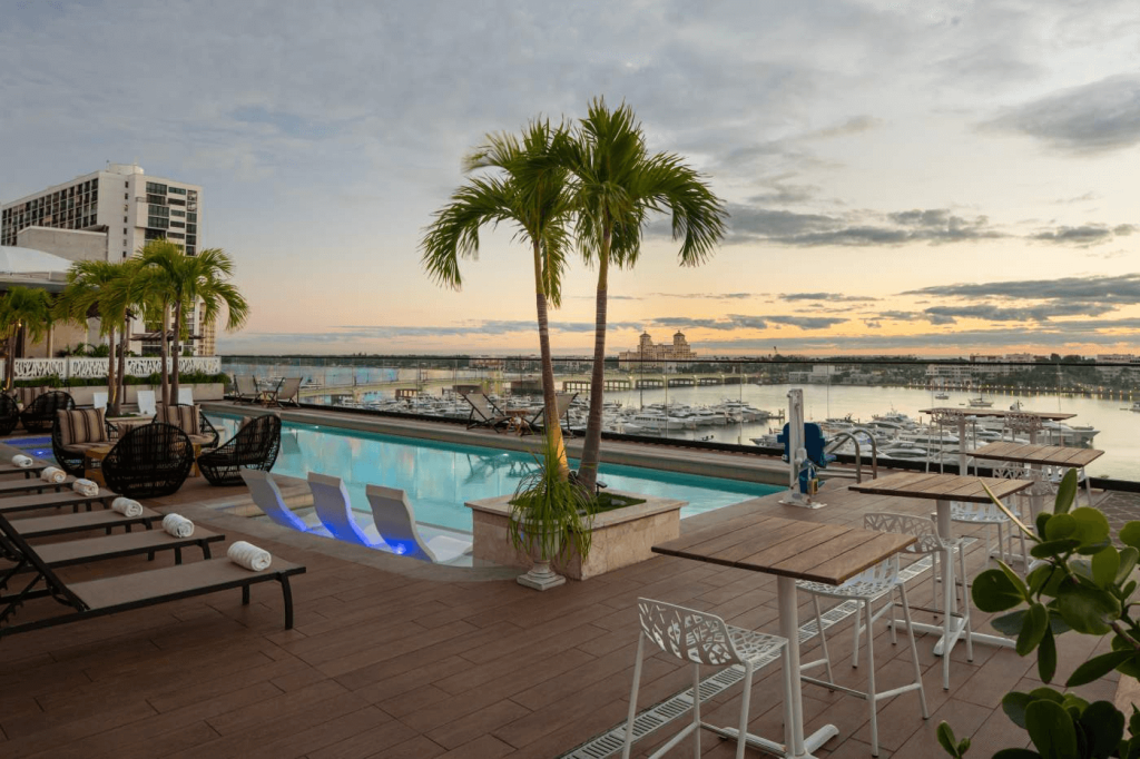 Uma área de piscina na cobertura com palmeiras, espreguiçadeiras e mesas com vista para o horizonte da cidade e a marina ao pôr do sol