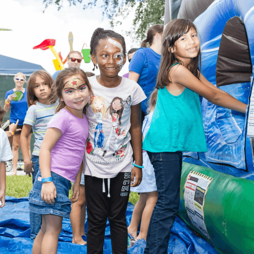 Three young girls with colorful face paint are standing in front of an inflatable bounce house at an outdoor event