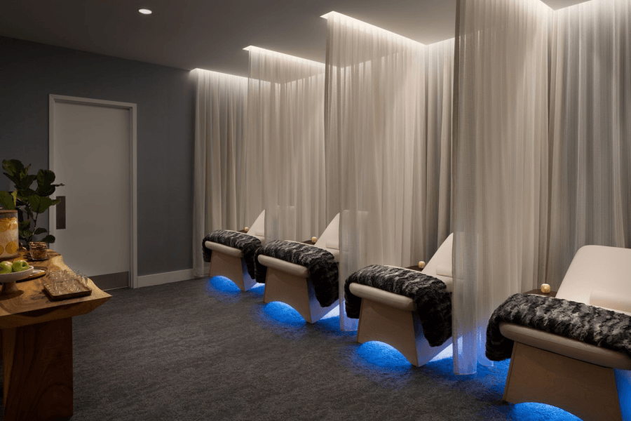 A serene spa room with four reclining chairs draped in sheer curtains, illuminated by soft blue lighting, and a wooden table with fresh fruit and a plant