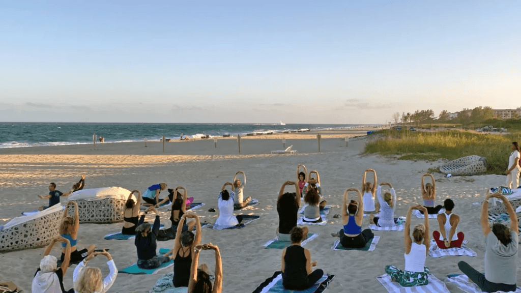 A large group of people practicing yoga on a serene beach at sunset, with the ocean waves in the background
