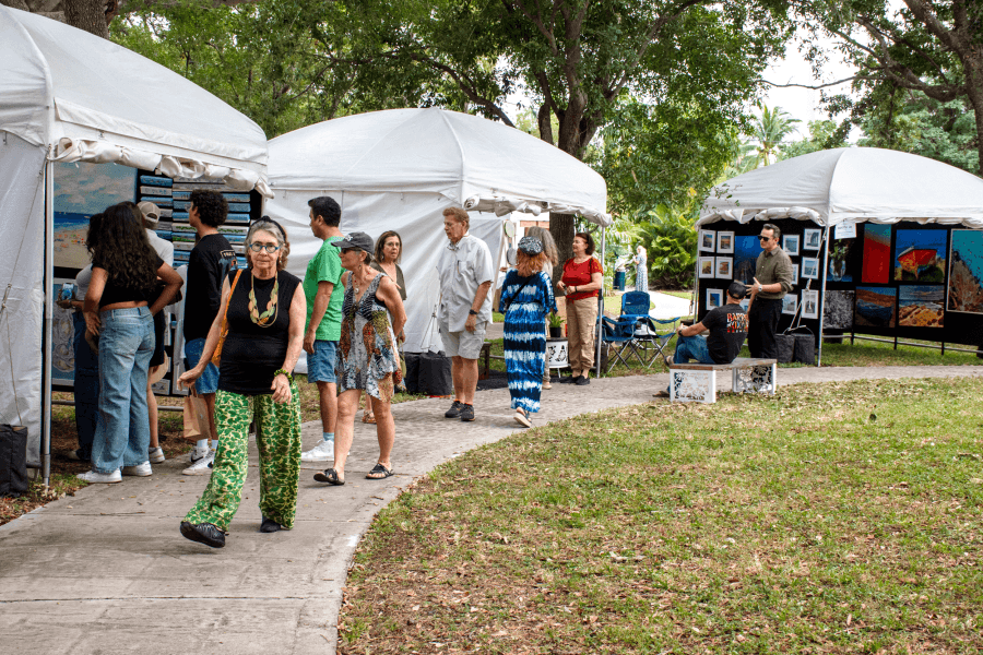 People stroll along a grassy path between white tents at an outdoor art fair, browsing paintings and prints displayed on easels and walls