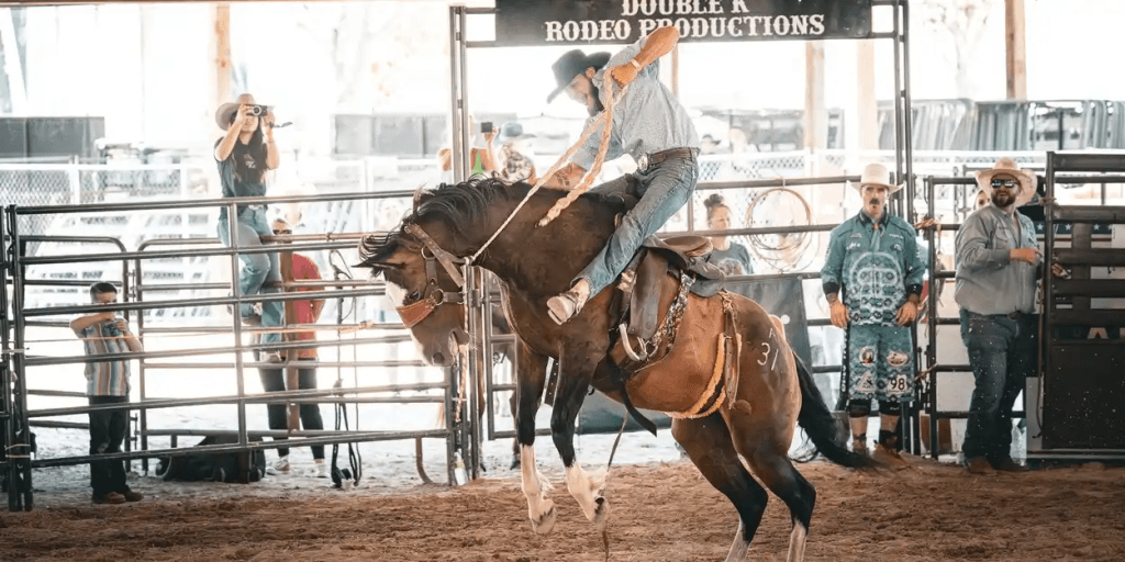 A cowboy in a white shirt and cowboy hat skillfully rides a bucking bronco at a rodeo, with spectators and event staff watching from the sidelines
