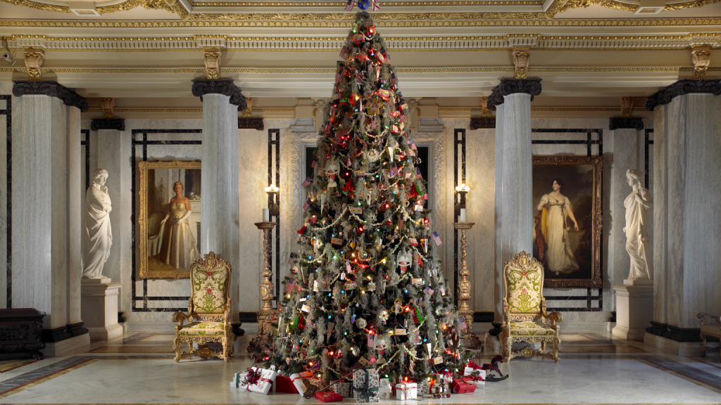 Un imponente árbol de Navidad se alza en una sala ornamentada con columnas de mármol, adornada con decoraciones elaboradas y rodeada de regalos