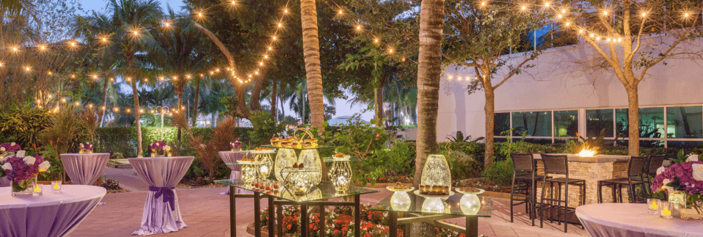 An elegant outdoor evening event with string lights, purple tablecloths, and a variety of desserts and drinks