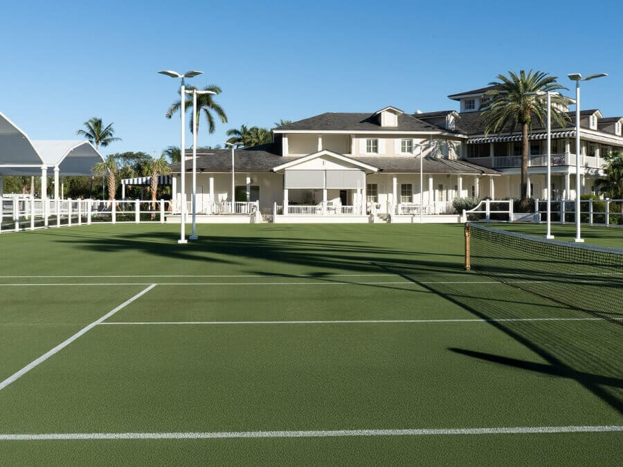 A well-maintained tennis court with a net, surrounded by palm trees and a white house with a porch