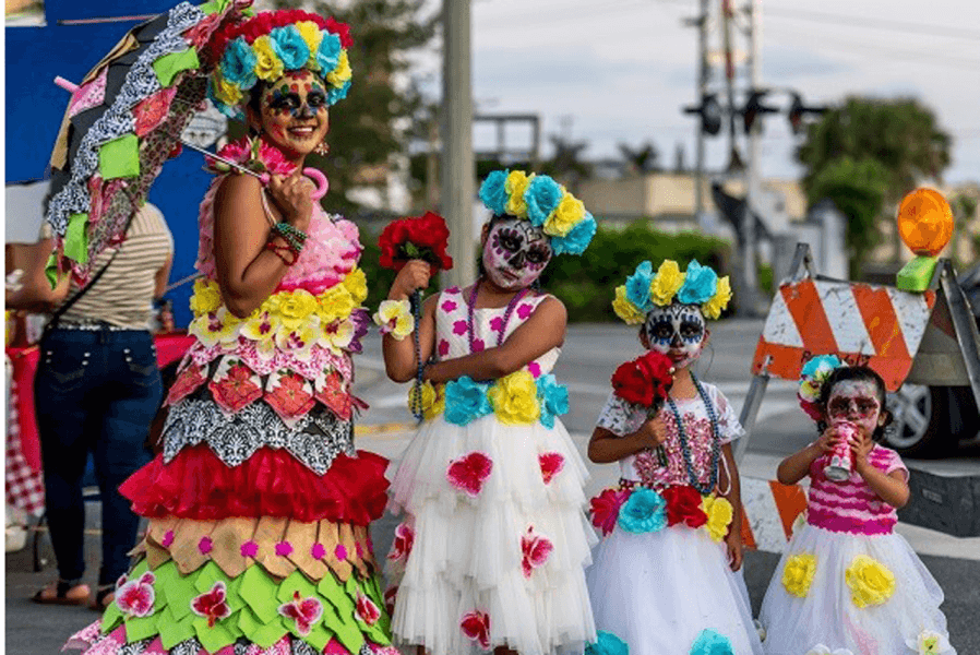 Four women dressed in vibrant, layered costumes adorned with colorful flowers and holding umbrellas celebrate a festive occasion