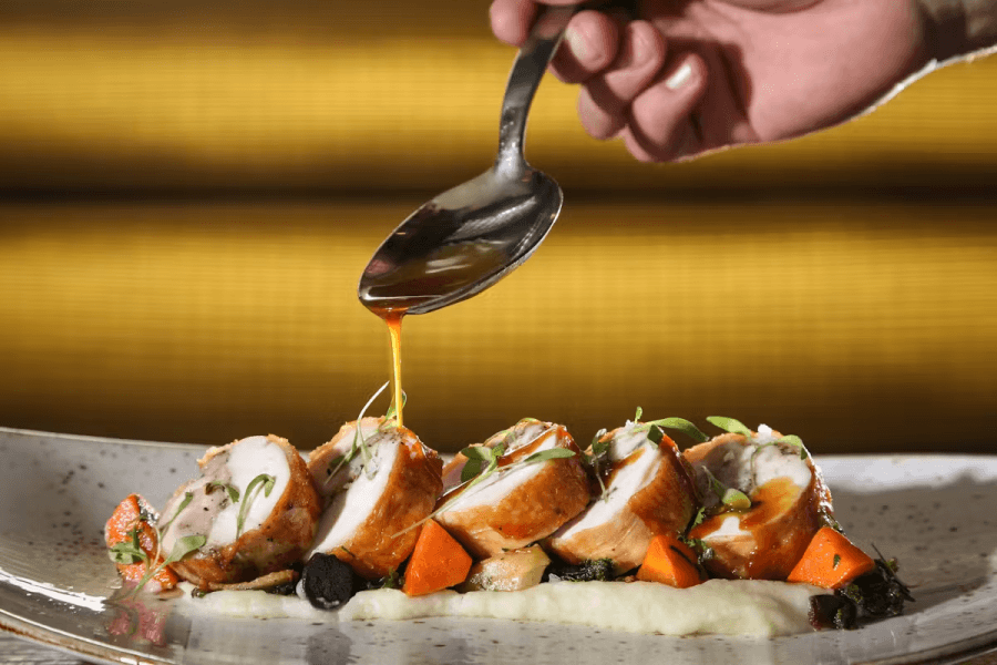 A hand pours a rich, golden sauce over slices of roasted chicken, garnished with fresh herbs and served on a bed of mashed potatoes with roasted vegetables