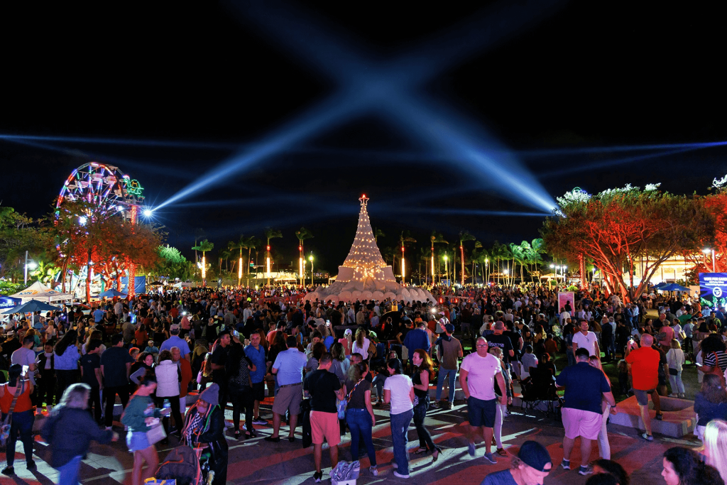 Una escena nocturna festiva en un evento navideño, con una gran multitud reunida alrededor de un imponente árbol de Navidad iluminado con luces de colores, acompañado de una colorida noria y palmeras bañadas en luz azul