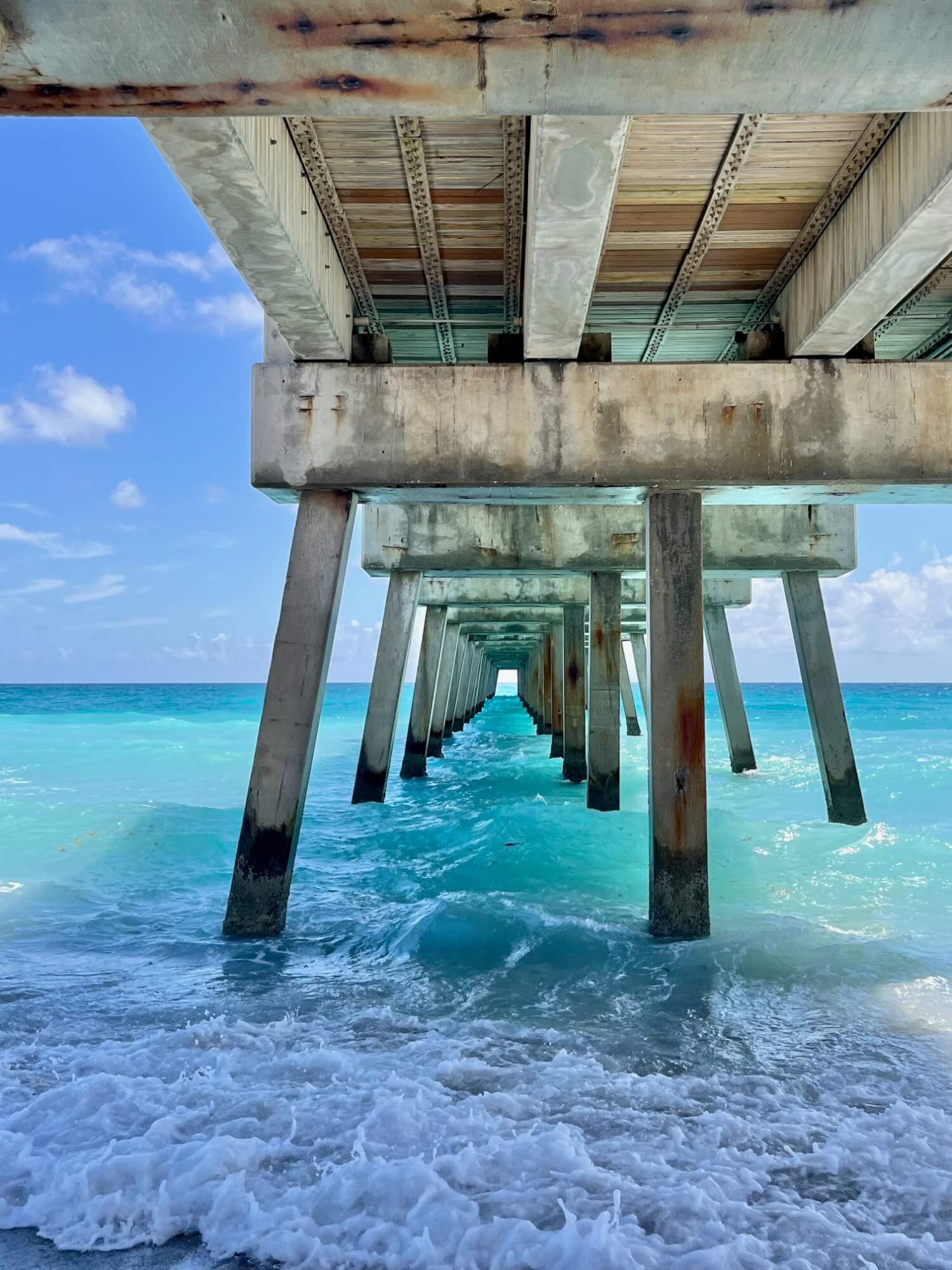 A long wooden pier extends into the turquoise ocean, supported by sturdy concrete pillars, with waves crashing beneath it against a backdrop of a clear blue sky