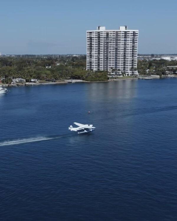 A small seaplane skims across calm waters near a city skyline