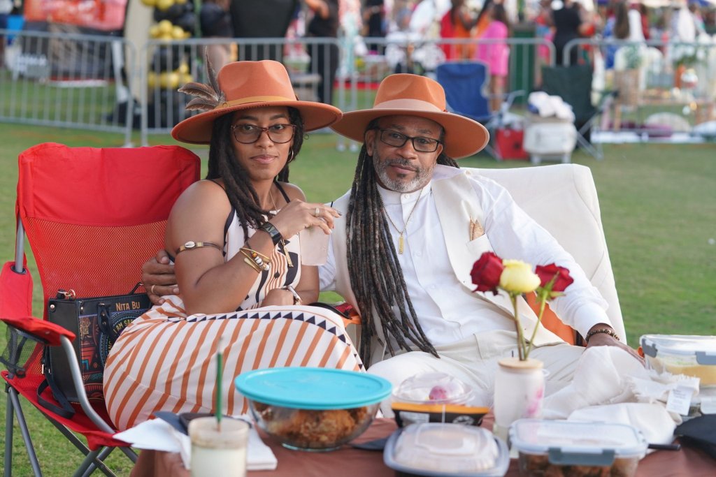 A couple wearing matching orange hats and white outfits sit together at a picnic table with food and drinks, surrounded by a crowd at an outdoor event