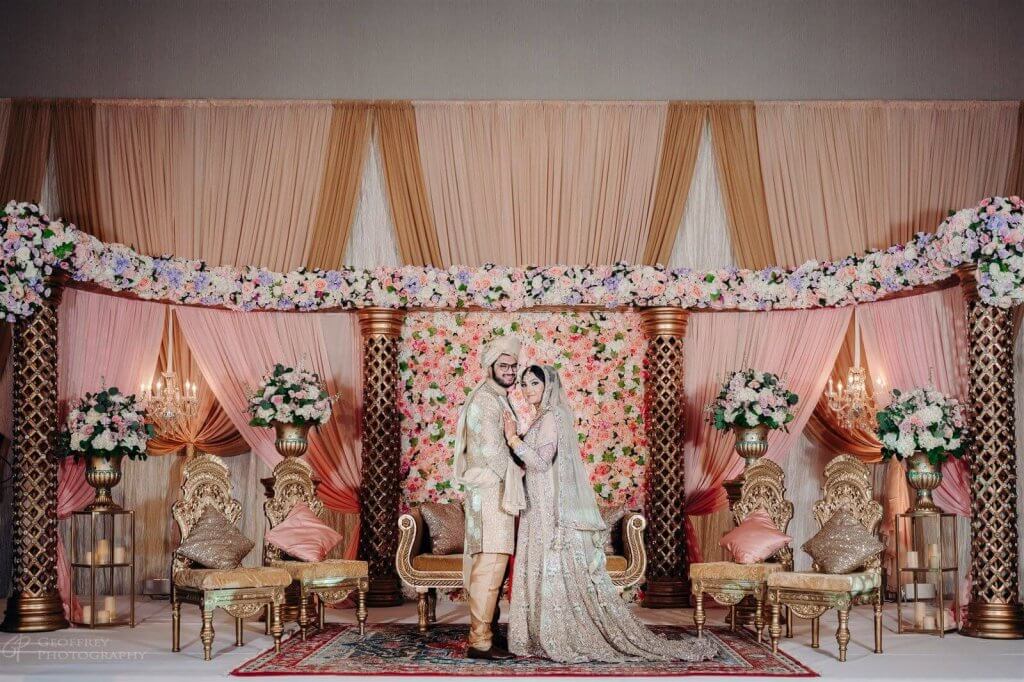 A lavishly decorated wedding stage with a floral archway, ornate chairs, and a golden throne-like sofa