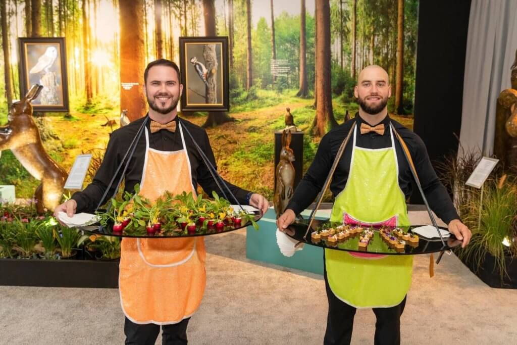 Dois homens de avental estão em uma sala com tema de floresta, segurando bandejas de alimentos e bebidas