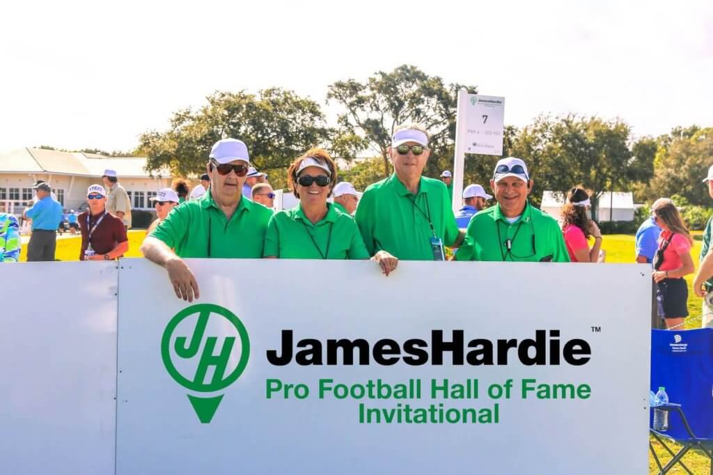 Eine Gruppe von fünf Personen mit grünen Hemden und weißen Mützen steht hinter einem Schild für das JamesHardie Pro Football Hall of Fame Invitational Golfturnier