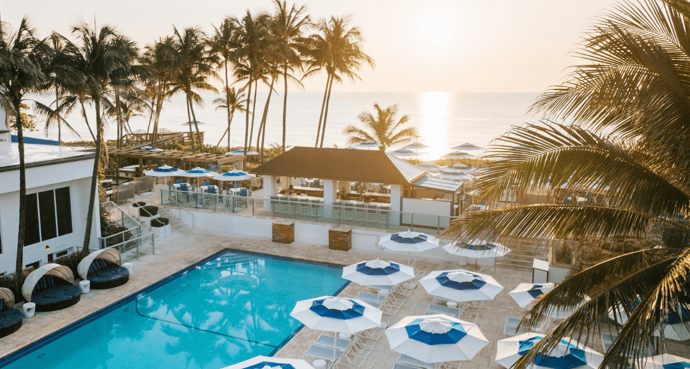 A serene beachfront resort with a large swimming pool surrounded by palm trees and white umbrellas, overlooking the ocean at sunset