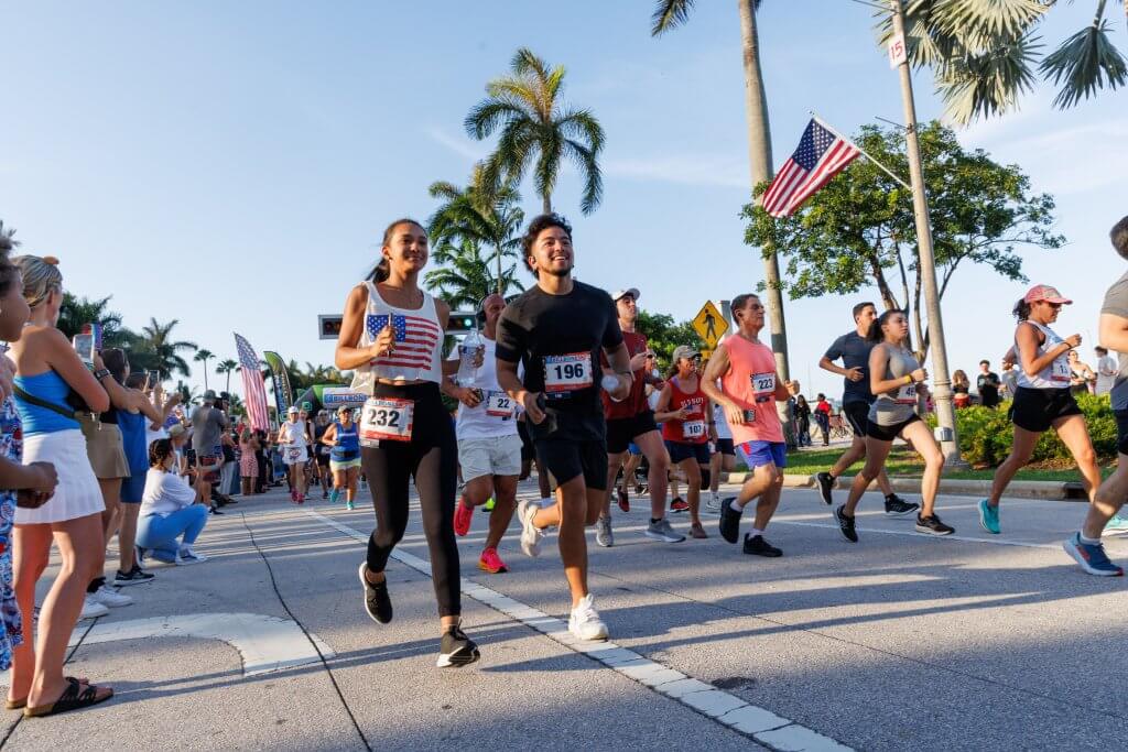 A diverse group of runners, including a man and woman in the lead, participate in a marathon with spectators cheering them on along a palm-lined street