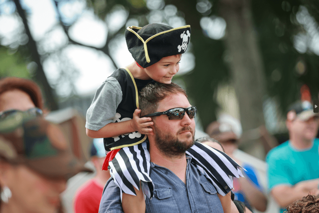 A young boy dressed as a pirate sits on his father's shoulders, both smiling and enjoying a sunny day outdoors