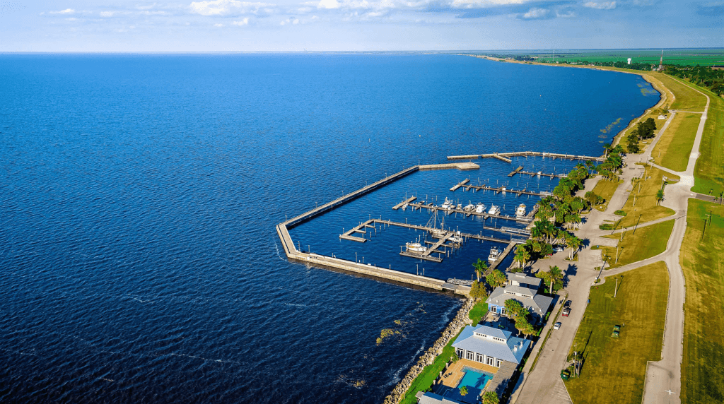 Aerial view of a marina with multiple docks and boats, adjacent to a sandy beach and a residential area with houses and palm trees