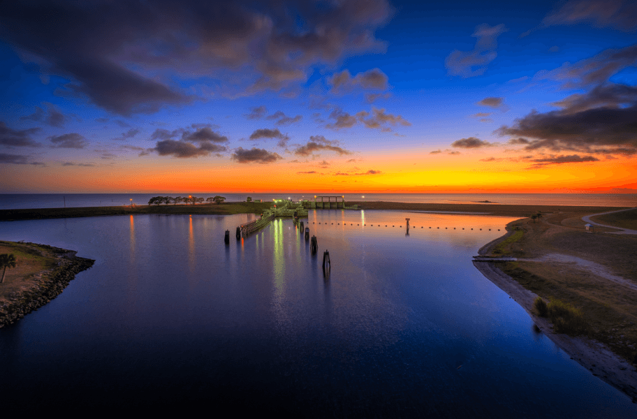 Coucher de soleil serein sur une étendue d'eau calme avec une jetée s'étendant au loin, reflétant des teintes vibrantes d'orange, de rose et de violet dans le ciel.