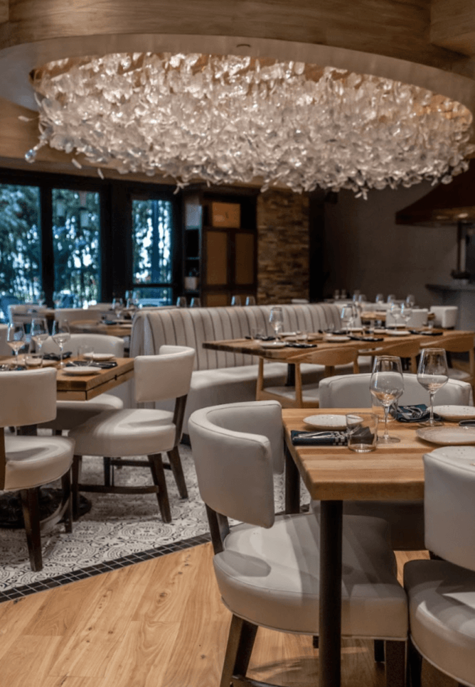A modern restaurant interior with a stunning chandelier, white leather chairs, and wooden tables set for dining