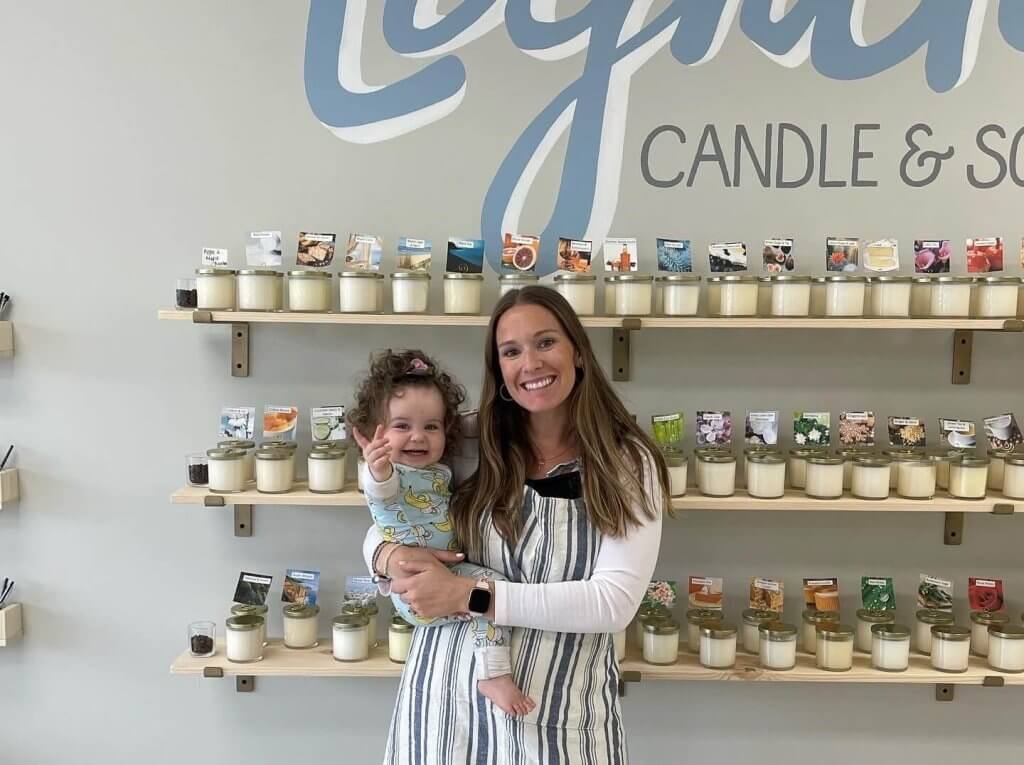 A smiling woman in a striped apron holds a happy baby in her arms, standing in front of shelves filled with neatly arranged scented candles in a cozy candle shop