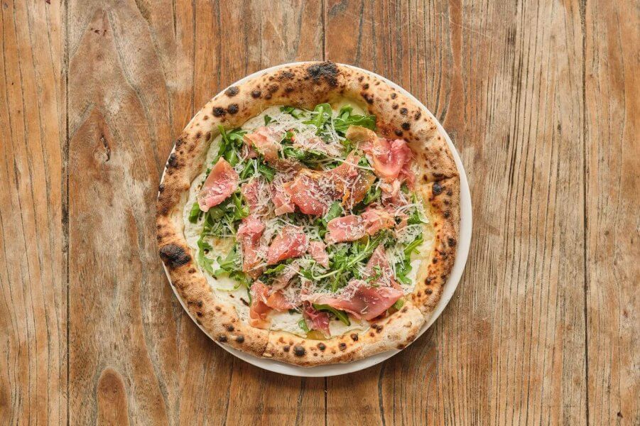 A rustic pizza with a charred crust, topped with fresh arugula, prosciutto, and shaved Parmesan cheese