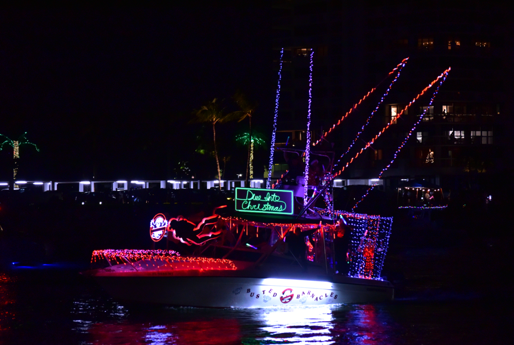 Ein festlich geschmücktes Boot mit leuchtenden Lichtern und einem "Dive into Christmas"-Schild nimmt an einer nächtlichen Bootsparade teil
