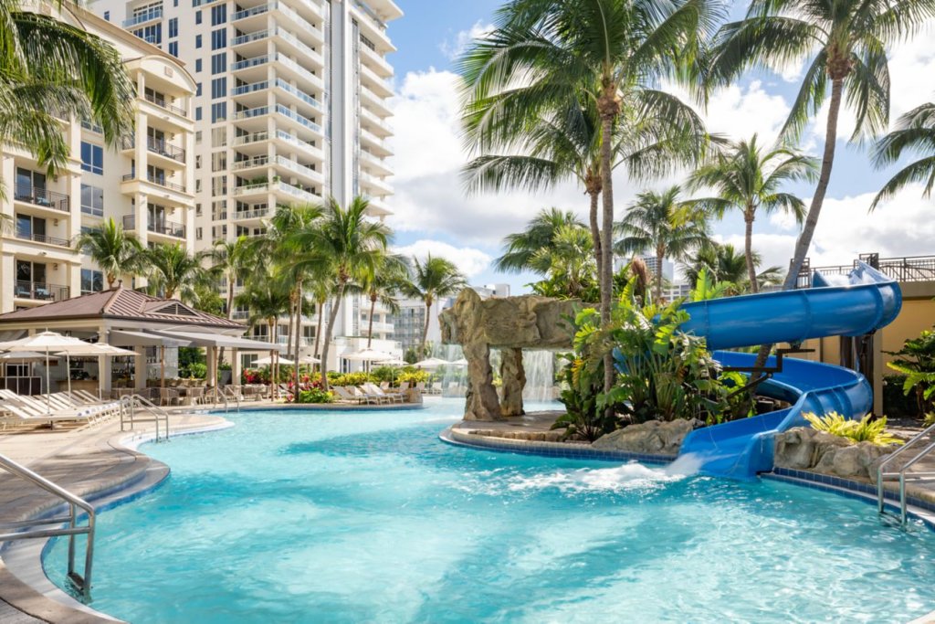 Ein lebhaftes tropisches Resort mit einem großen, nierenförmigen Swimmingpool mit einer blauen Wasserrutsche, die sich durch üppiges Grün und Palmen schlängelt, umgeben von Hochhäusern