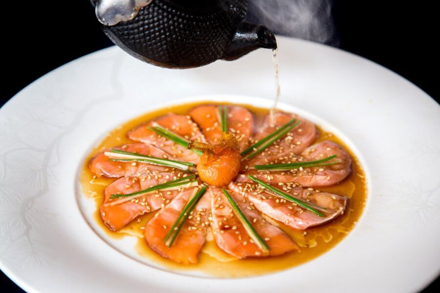 A delicate dish of thinly sliced salmon arranged in a circular pattern on a white plate, garnished with sesame seeds and green onions, with a black teapot pouring a dark sauce over it