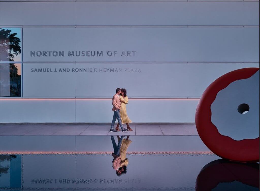 Un couple s'embrasse devant le Norton Museum of Art, avec une grande sculpture de beignet rouge à proximité.