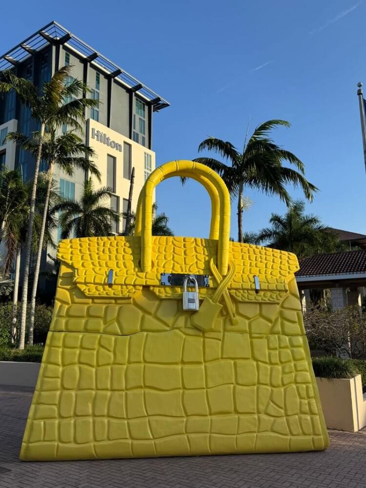 A giant yellow crocodile-skin handbag sculpture stands in front of a Hilton hotel, surrounded by palm trees